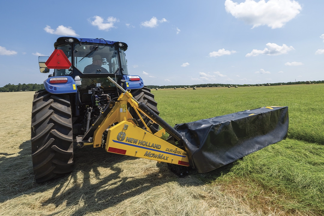 Tractor azul operando segadora DuraDisc 109M en campo con pacas de heno.